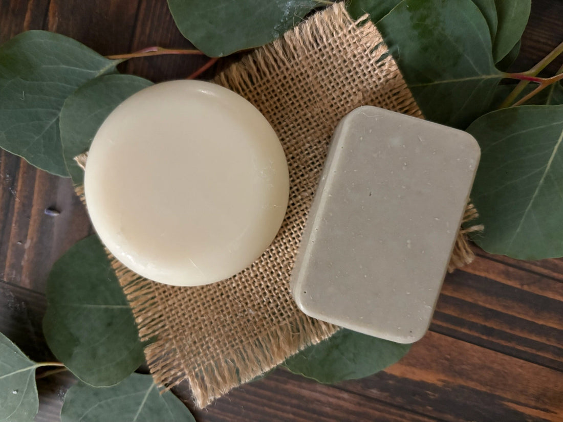 Two handcrafted bars of goat milk soap—one round and creamy white, the other rectangular and textured with natural botanical specks—resting on a rustic piece of burlap alongside fresh green eucalyptus leaves.